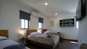 Quarto de hotel com três camas, uma pequena mesa, uma cadeira e uma televisão.