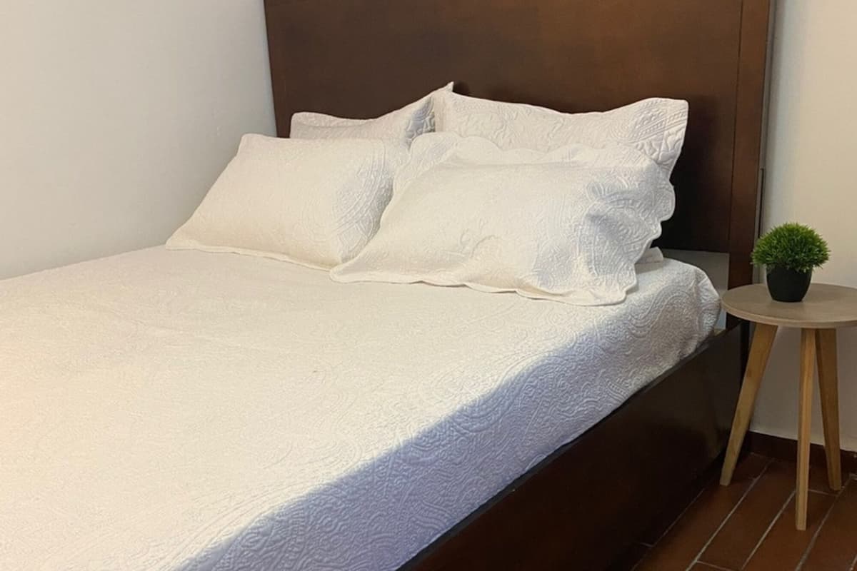 Un llit amb un capçal de fusta, roba de llit blanca i una petita planta en test amb una taula rodona de fusta al costat.