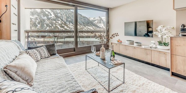 A modern living room with a large window, a sofa, a coffee table, and a kitchen area.