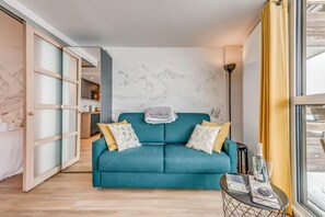 A teal sofa with two pillows, a small table with a glass and a bottle, and a wall with a mountain design.