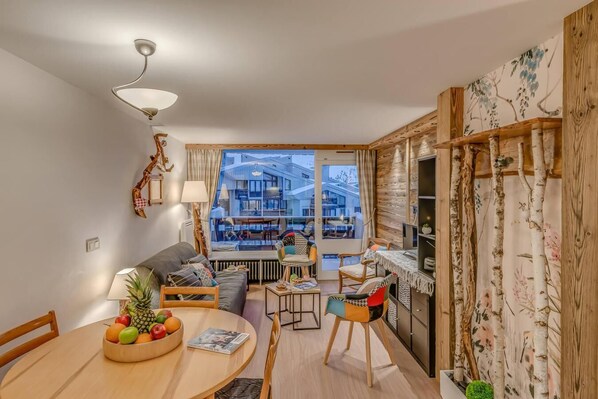 A modern living room with a sofa, dining table, and chairs, a view of a building through a glass door, and wooden decor. - 300m from resort center, 4th floor, balcony, tv, ski locker, 39m², Tignes (Tignes)