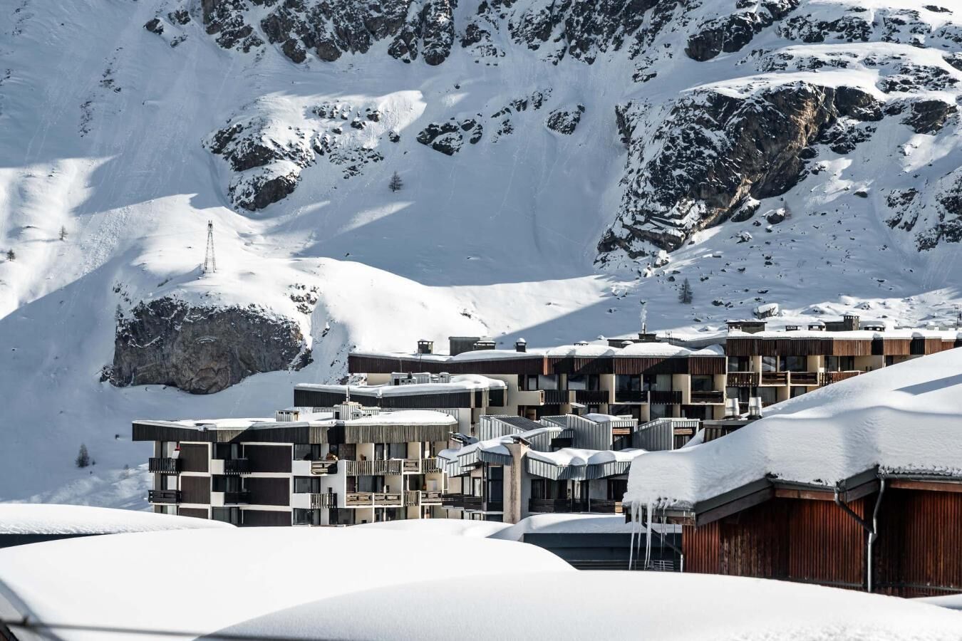 A snow-covered mountain with modern buildings, including a multi-story structure with balconies and a wooden building with a sloped roof.