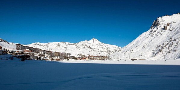 Une station de montagne enneigée, avec un vaste ensemble de bâtiments, des pistes de ski et un ciel d’un bleu éclatant.