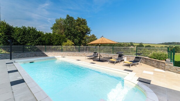Una piscina rectangular con borde de piedra, rodeada por una valla metálica y equipada con sillas de descanso y sombrillas.