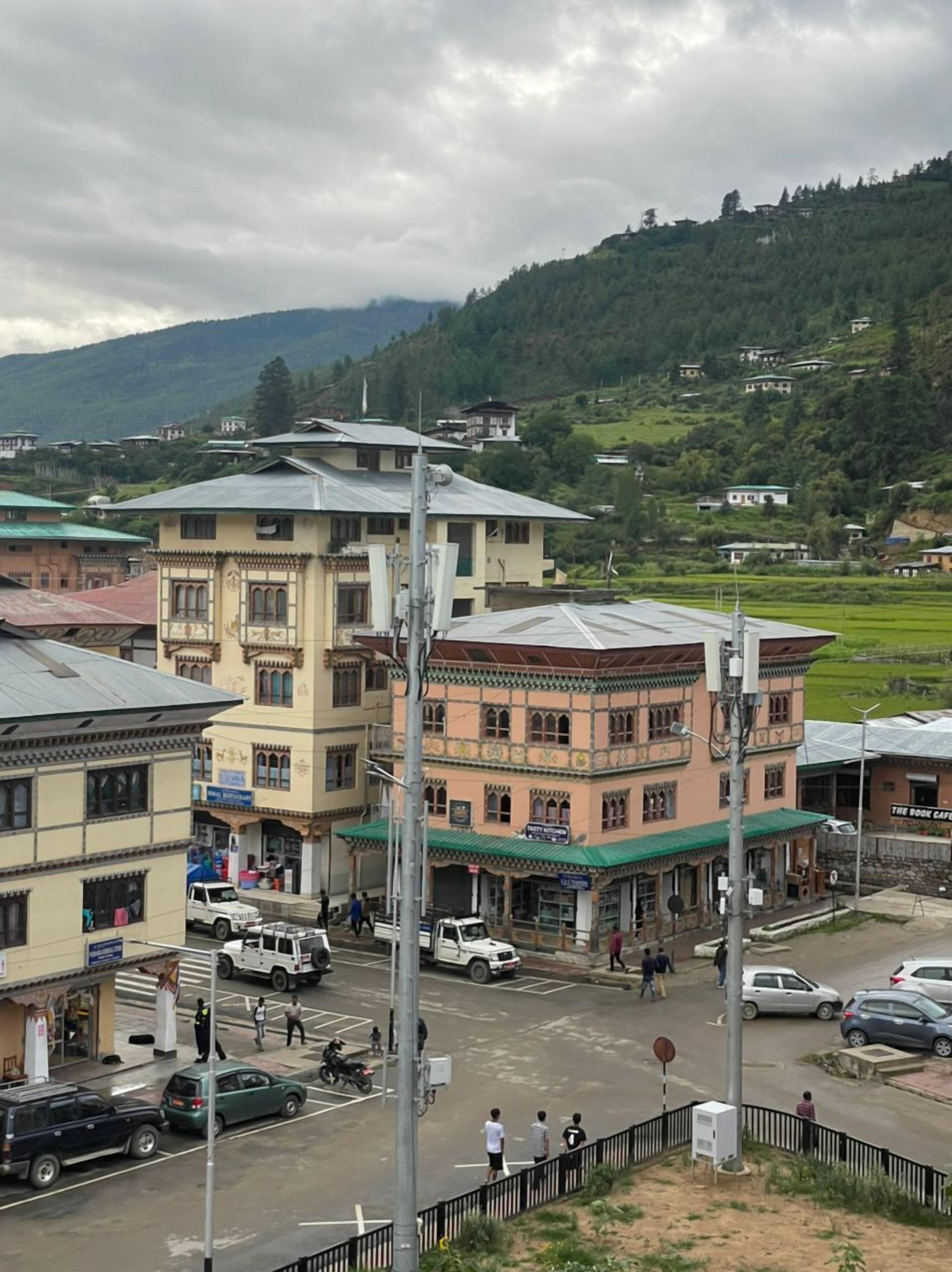 A multi-story building with traditional architecture, a street in front with parked cars, and a hilly landscape in the background.