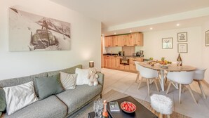 Una sala de estar moderna con un sofá gris, una zona de comedor con una mesa redonda y sillas, y una cocina al fondo.