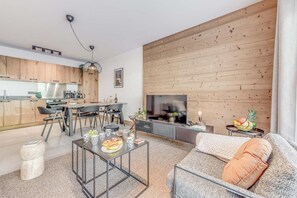 A modern living room with a sofa, coffee table, and TV. There is a kitchen area with wooden cabinets and a dining table.