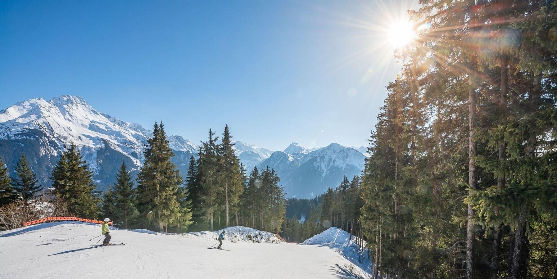 A snowy ski slope with skiers, surrounded by tall evergreen trees and snow-covered mountains under a clear sky.