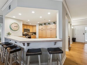 Una cocina moderna con una isla central, taburetes de bar, armarios de madera, un microondas y un reloj en la pared.