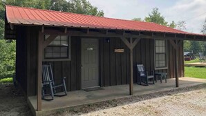 A rustic cabin with a red metal roof, a porch with two rocking chairs, and a small table. - Cozy and centrally located in the middle of all the best of nature’s offerings!  (Fifty-Six)
