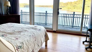 A bedroom with a large bed, a nightstand, a lamp, and a view of a lake through a sliding glass door. - Beautiful Oceanfront House- Stunning Water Views (Harpswell)