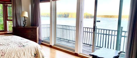 A bedroom with a bed, a nightstand, a lamp, and a view of a lake through large windows.
