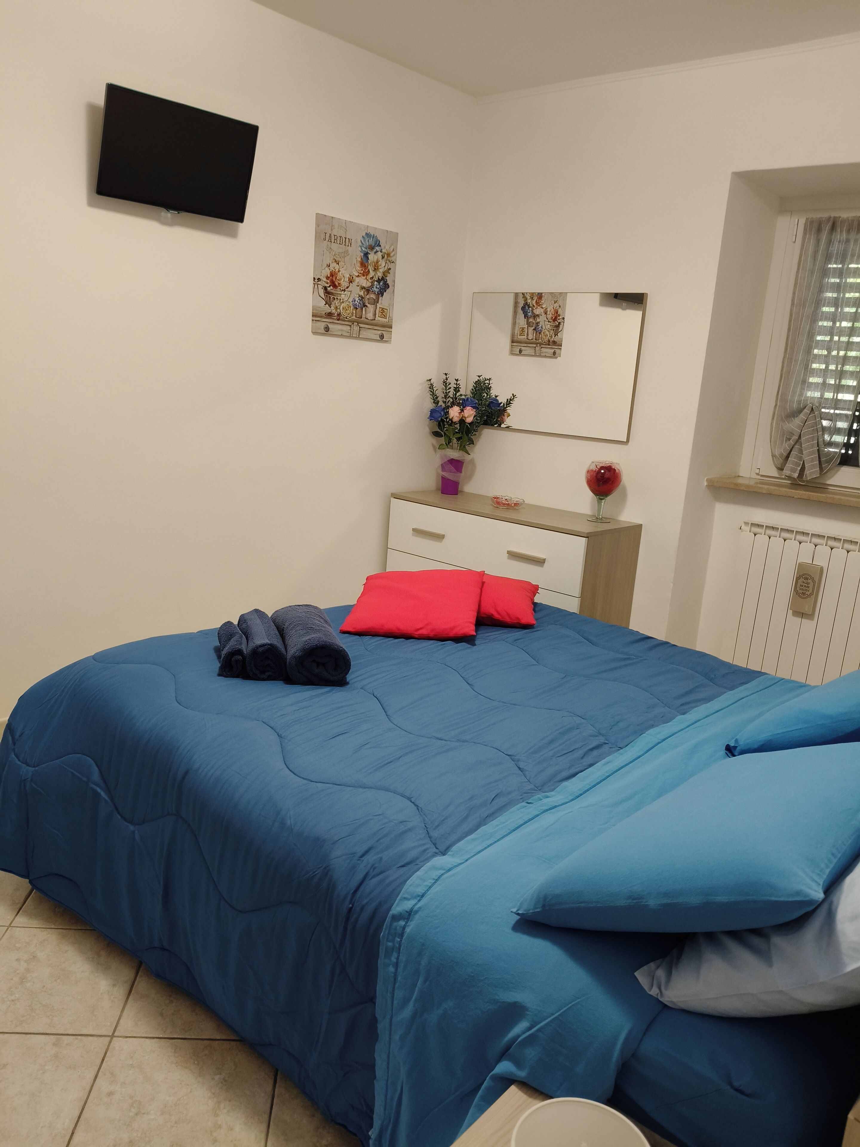 Ein Schlafzimmer mit blauer Bettdecke, roten Kissen, einem aufgerollten Handtuch, einem Schminktisch mit Spiegel, einer Vase mit Blumen und einem Fernseher an der Wand.