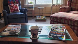 Un salon chaleureux avec une grande fenêtre donnant sur la mer, une table basse en verre sur laquelle reposent des magazines et une tasse, et deux fauteuils agrémentés de coussins colorés.