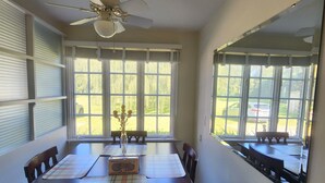 A dining room with a large table, chairs, a ceiling fan, and large windows with blinds.