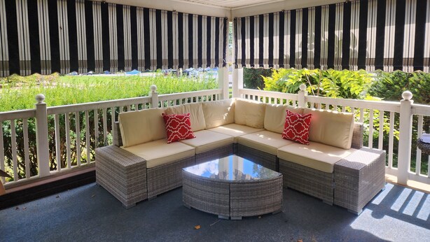 A covered outdoor seating area with a white railing, a wicker sofa with cushions, and a glass coffee table.