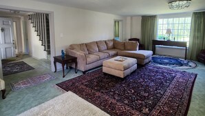 A spacious living room with a beige sectional sofa, a matching ottoman, and a large patterned rug. A staircase is visible in the background, leading to another level.