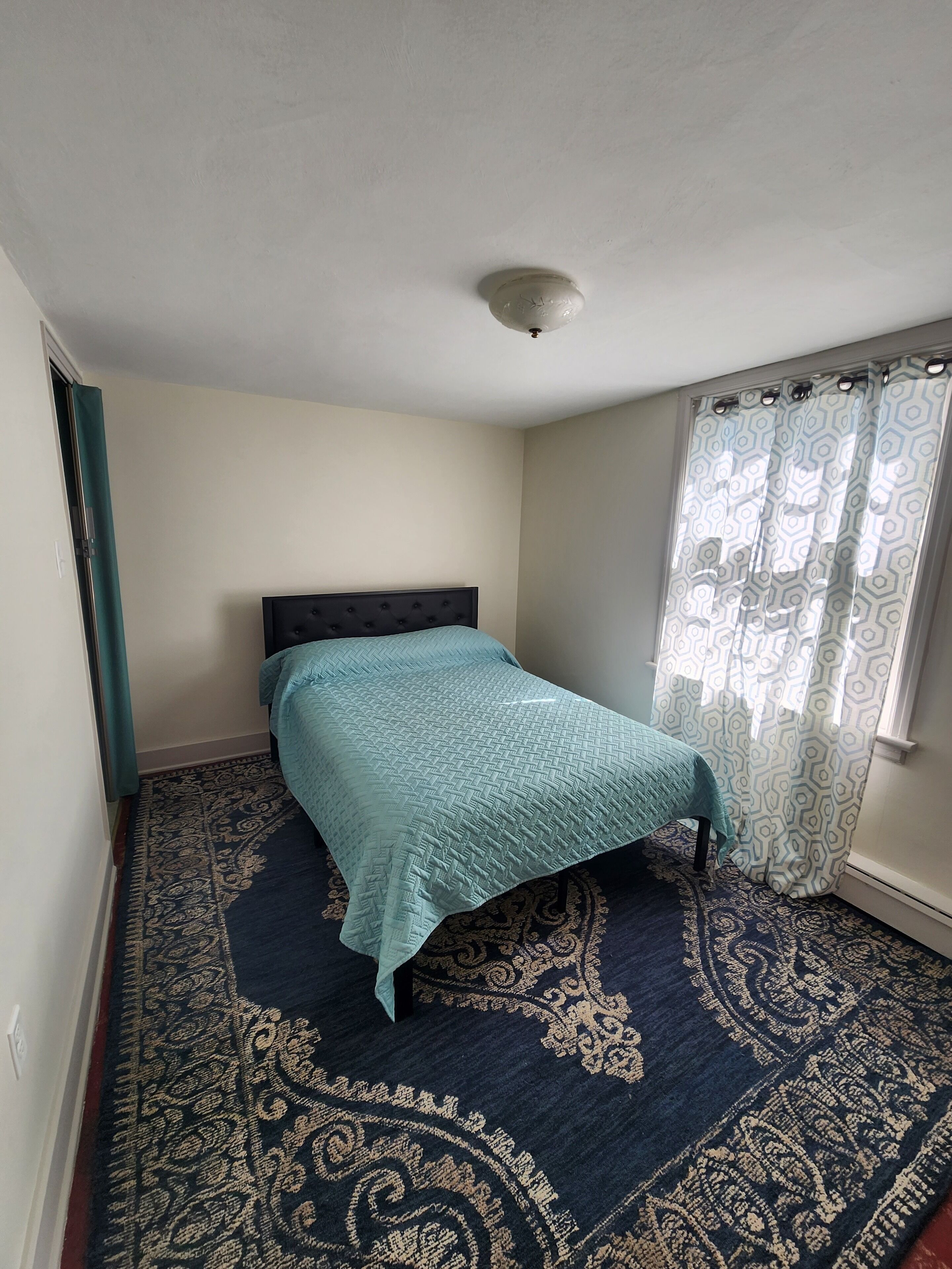 A bedroom with a bed, a patterned carpet, a window with curtains, and a ceiling light.
