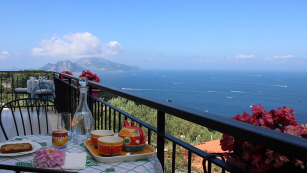 Un balcone con un tavolo apparecchiato per la colazione, vista mare, con una bottiglia, piatti e un fiore.
