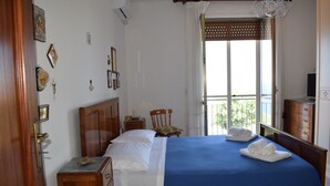 Un letto rifatto con una coperta blu, una testiera in legno e un comodino con una lampada. Si vede una finestra con vista su del verde all'esterno.