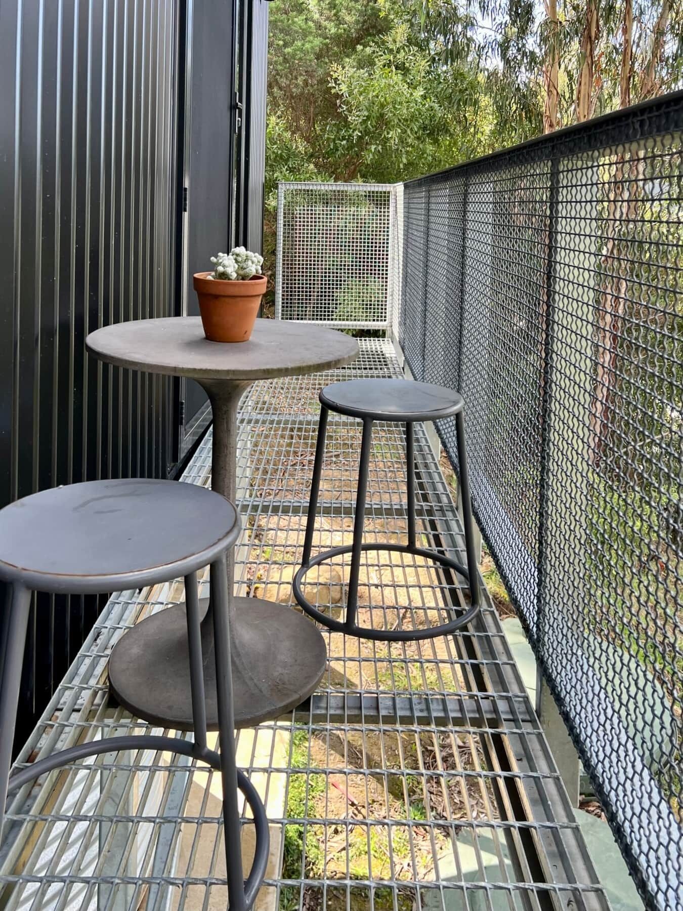Parvekkeella on pyöreä pöytä ja kaksi jakkaraa, ja se on ympäröity metallisella aidalla sekä koristekasvilla, joka on istutettu ruukkuun.