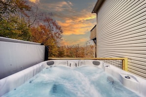 Een hottub met een houten terras, omringd door bomen en een gebouw met horizontale beplating.