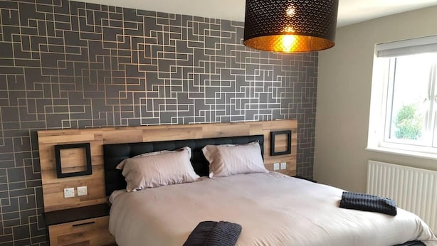 A modern bedroom with a geometric-patterned wall, a wooden headboard, a black upholstered bed, and a hanging light fixture. - 3 Bed House - Sleeps 6 - Garden - Parking - Wifi (Rye)