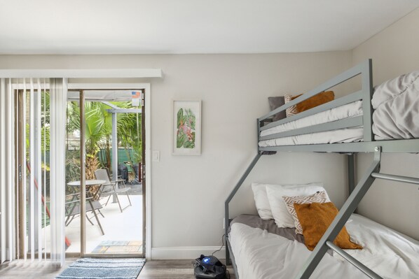 A bunk bed with white bedding and a brown pillow, a small table with chairs, and a sliding glass door leading to an outdoor area with a patio and chairs. - Wonderful 3bdr cottage, 5mins from beach (Jupiter)