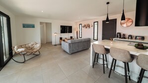 A modern kitchen with a bar area, a grey sofa, and a hanging light fixture. - Villa Azuréa Piscine et Plages (Sainte-Anne)