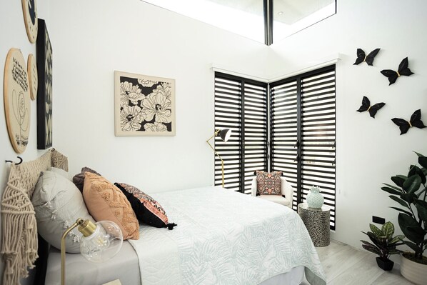 Une chambre à coucher comprenant un lit, des oreillers, une table de chevet, une lampe, une fenêtre avec des stores, des œuvres d’art murales et une plante.