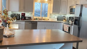 A modern kitchen with white cabinets, a large island with bar stools, a microwave, a refrigerator, and a window with a view of trees. - Huron waterfront cottage/Hot tub/max2 pets for16+people with private sand beach  (Goderich)