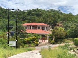 Una casa con techo rojo, rodeada de abundante vegetación y una colina al fondo.