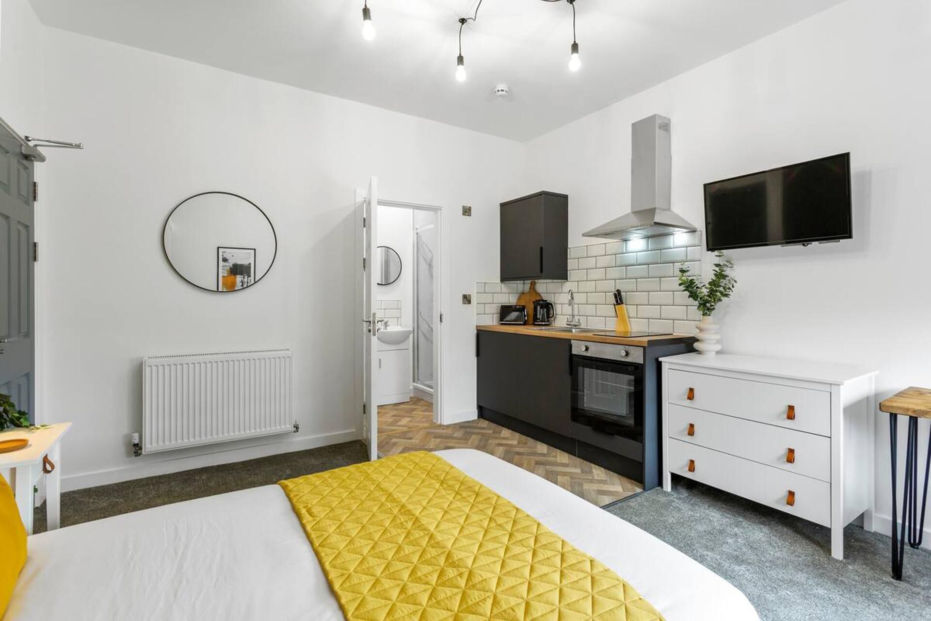 A modern hotel room with a bed, a bedside table, a round mirror, a white radiator, a kitchen area with a stove, a sink, and a TV mounted on the wall.