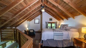 A bedroom with a sloped ceiling, wooden beams, a bed with pillows, a nightstand, and a window with curtains. - Hottest Sparrow - Charming Cottage (West End)