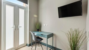 Une pièce avec un bureau de couleur turquoise, une chaise noire, une plante en pot, un téléviseur fixé au mur et une porte-fenêtre coulissante.