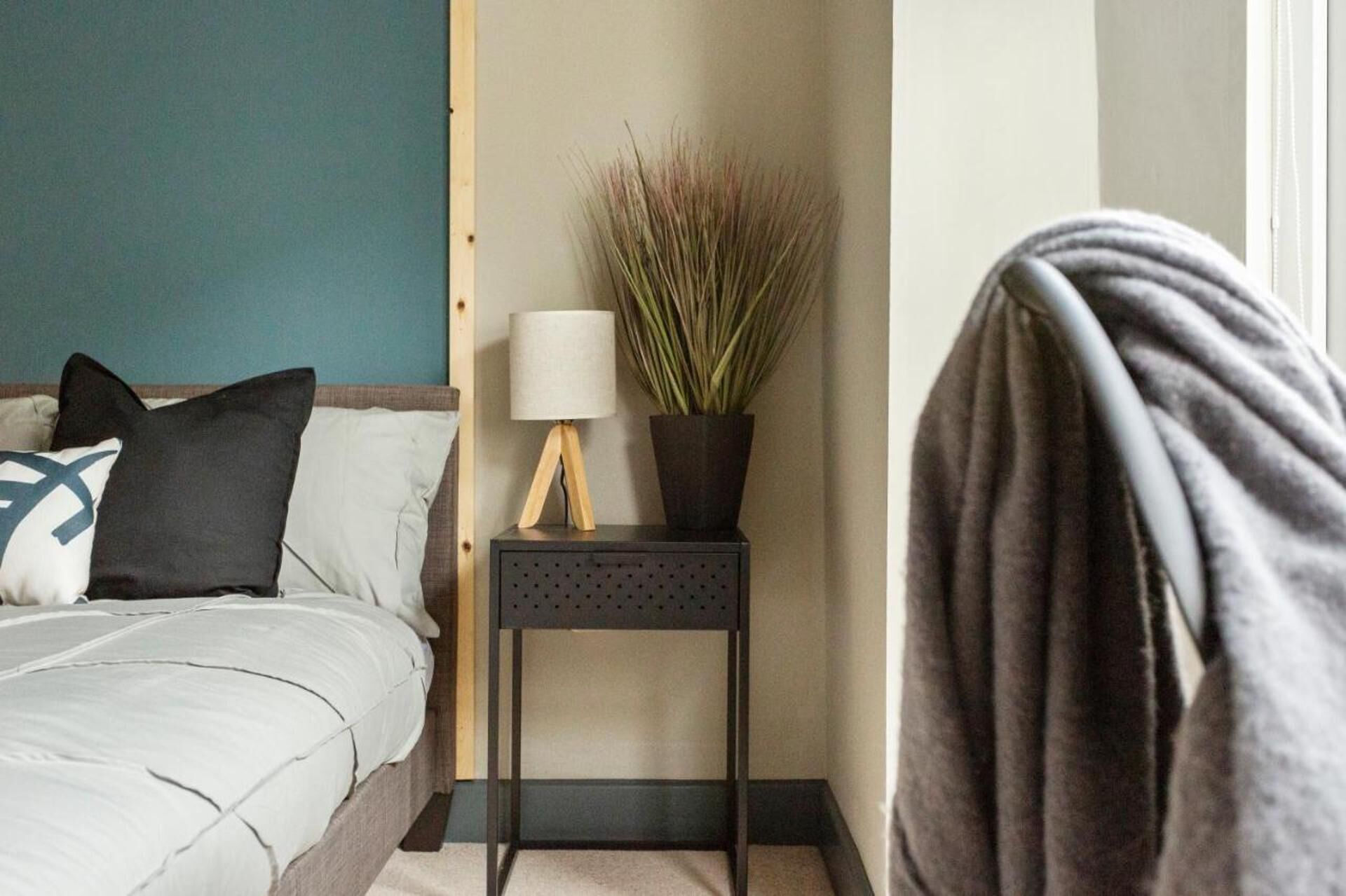 A bedroom with a bed, pillows, a bedside table with a lamp and a potted plant, and a blanket draped over the bed.