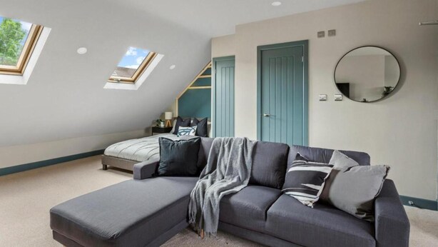 A modern bedroom with a large gray sectional sofa, a skylight, a bed with a blue headboard, and a round mirror on the wall.