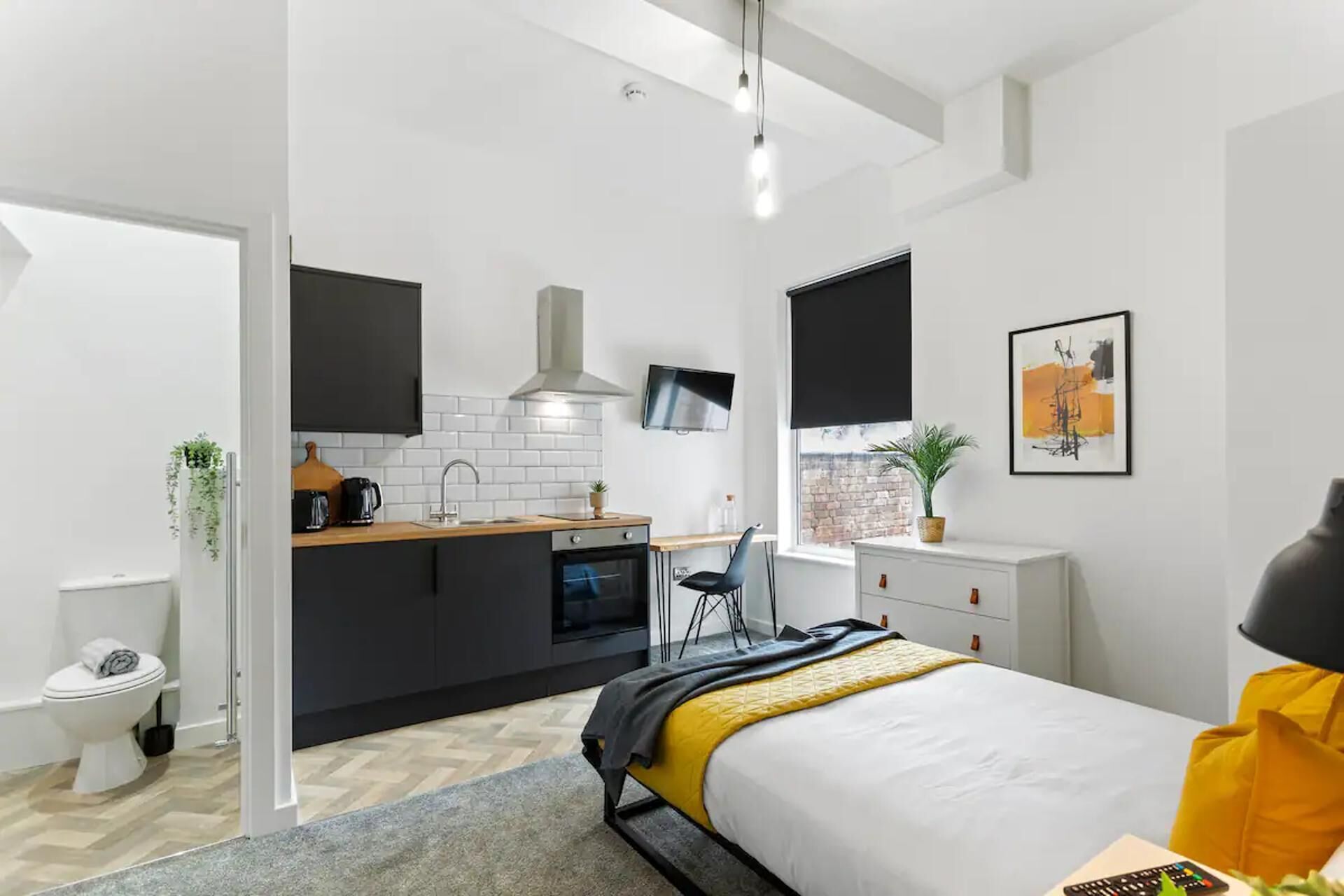A modern, compact bedroom with a bed, bedside table, and a window. Adjacent is a kitchenette with a sink, stove, and a TV.