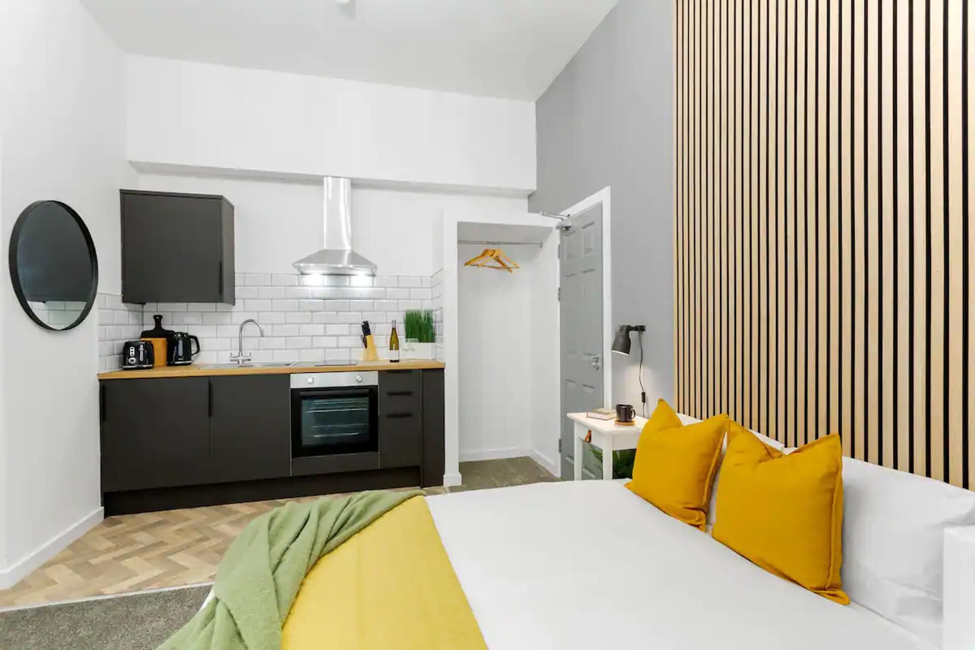 A compact hotel room with a kitchenette, a bed with yellow and white bedding, and a striped wall.