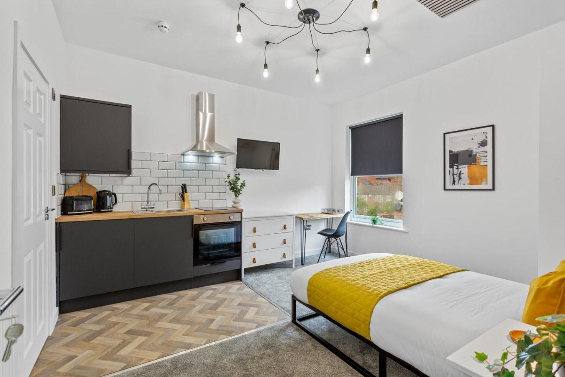 A modern, compact bedroom with a bed, bedside table, and a window. Adjacent is a kitchenette with a stove, sink, and a small dining area.