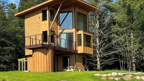 A modern wooden cabin with a balcony, surrounded by a grassy area and large rocks. - Enjoy whatever winter throws at you while you relax in the hot tub with a view! (Vermontville)