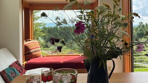 A room with a large window, a wooden table with a vase of flowers, and two glasses with apple designs. - Enjoy whatever winter throws at you while you relax in the hot tub with a view! (Vermontville)