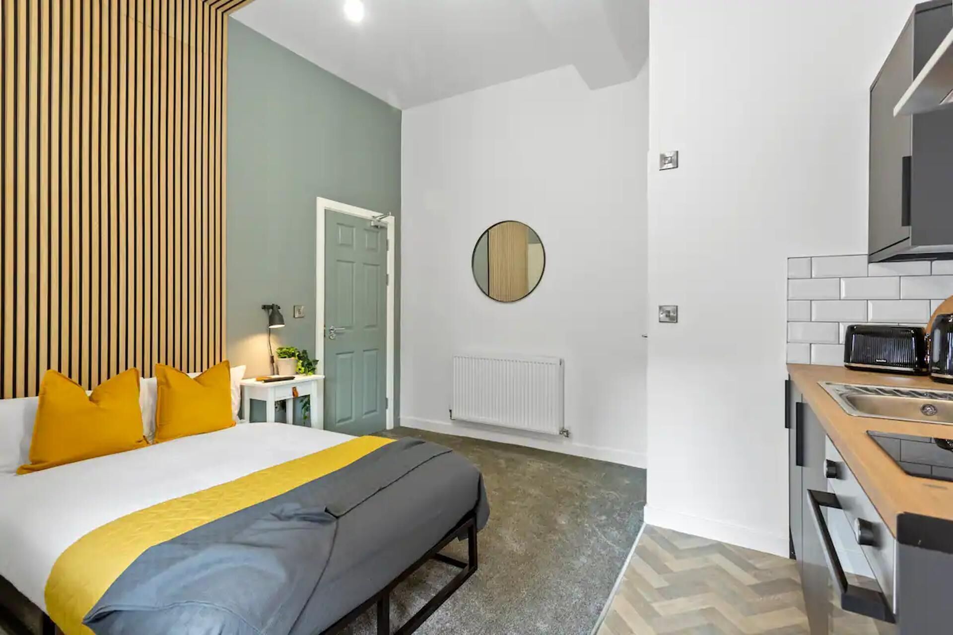 A modern hotel room with a bed, bedside table, lamp, and a round mirror on the wall.