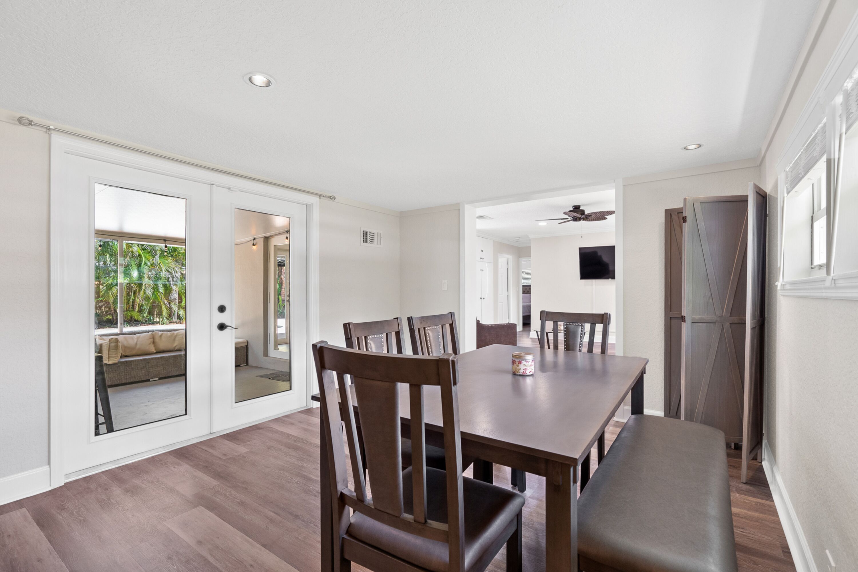 A dining area with a long table, multiple chairs, and a bench. There is a door leading to a balcony with a view of greenery.