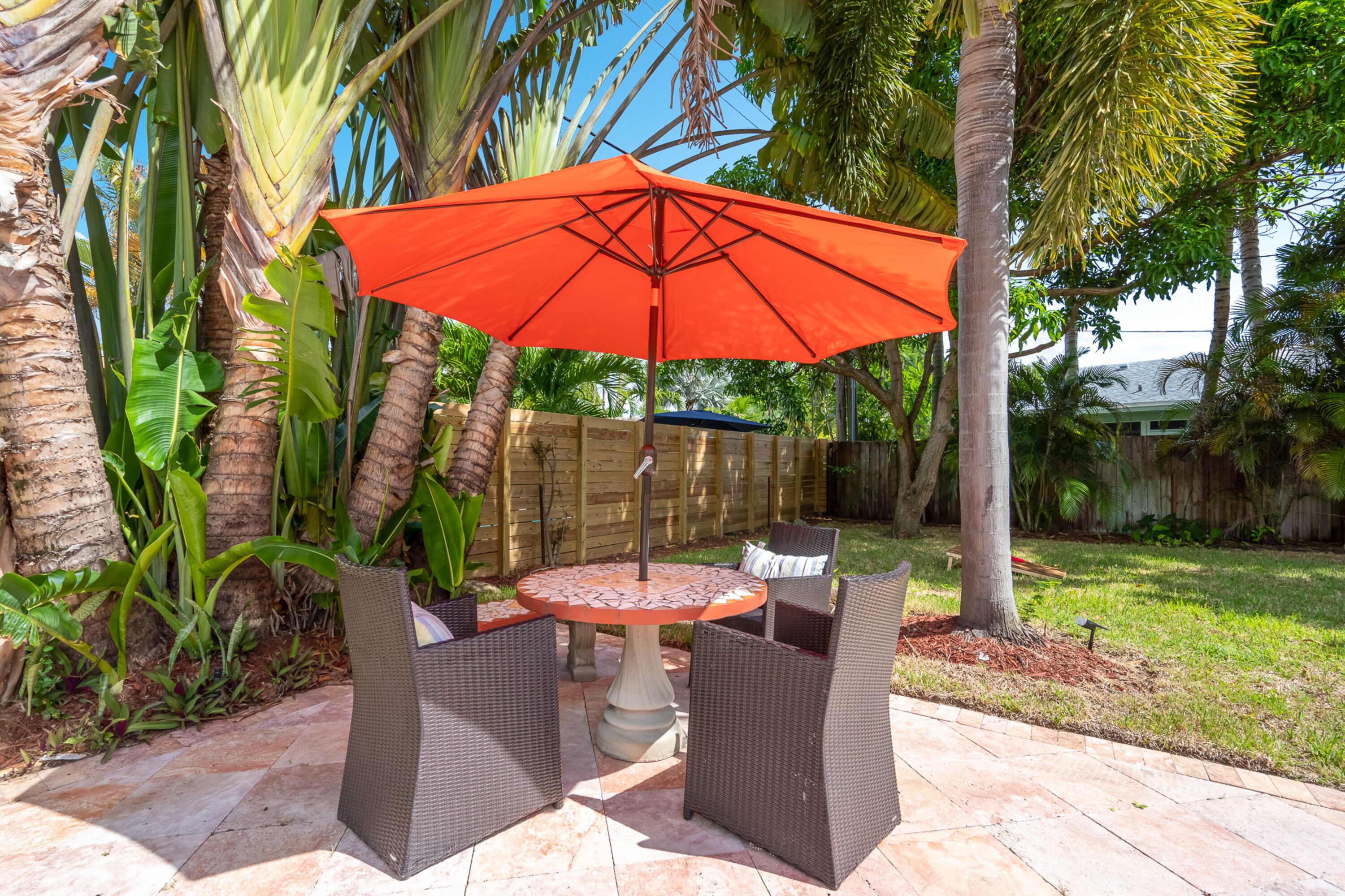 An outdoor patio with a round table and two chairs, an orange umbrella, and a wooden fence in the background.