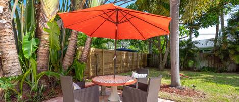 An outdoor patio with a round table and two chairs, an orange umbrella, and a wooden fence in the background.