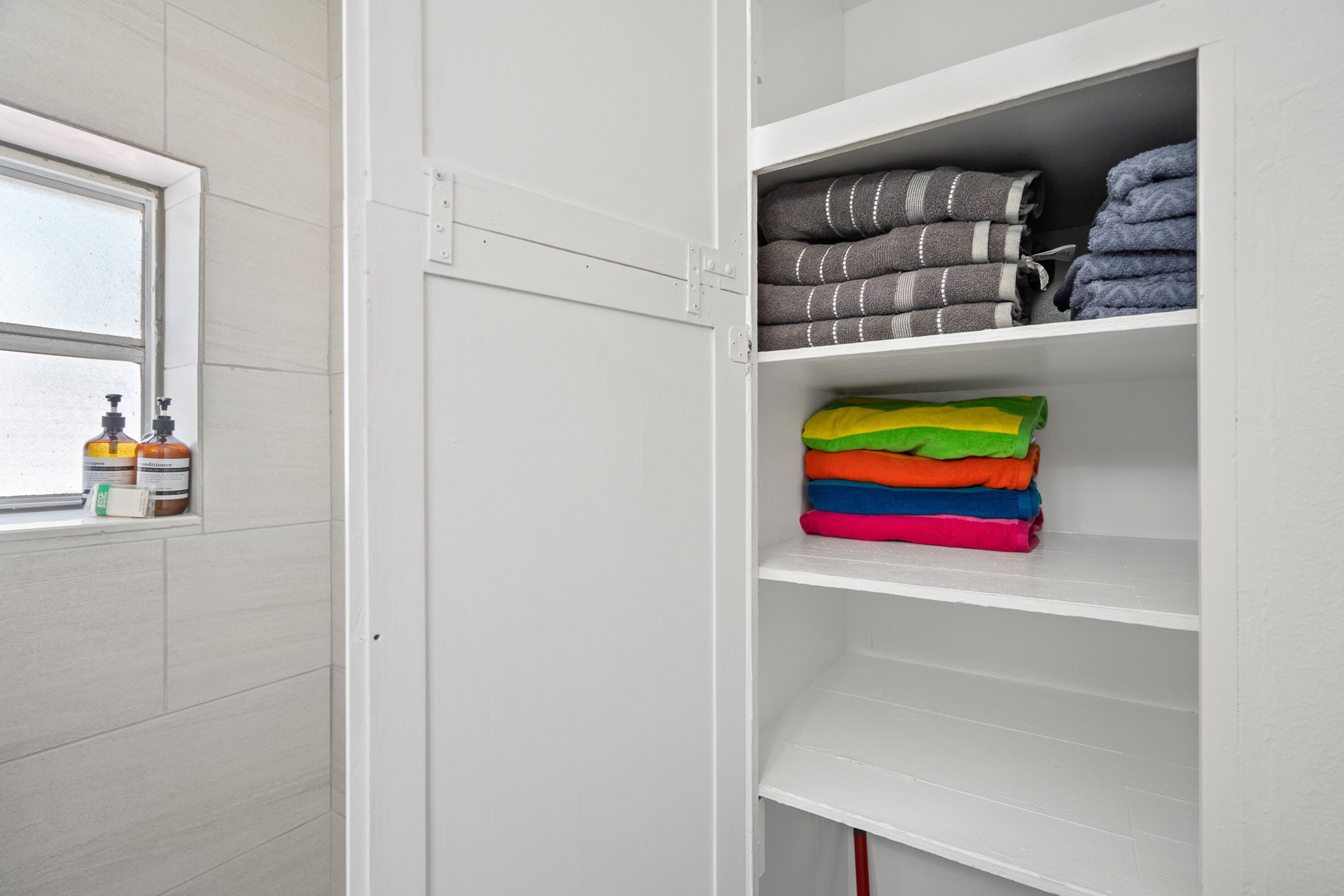 A white closet with shelves holding folded towels and a window with toiletries.