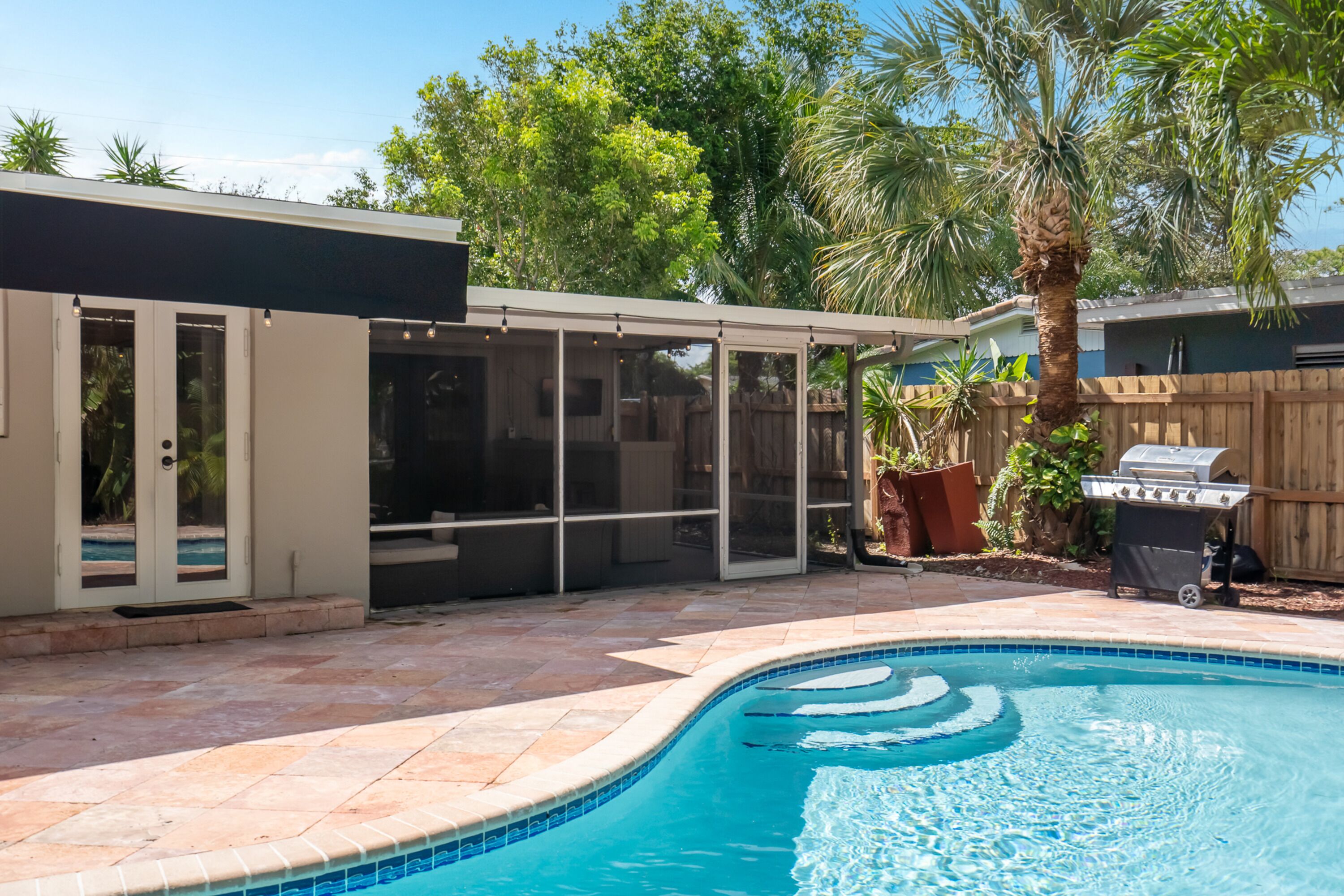 A swimming pool with a tiled edge, a covered patio area, and a wooden fence in the background.