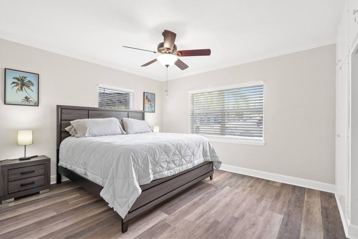 Une chambre à coucher comprenant un lit, une table de chevet, un ventilateur de plafond et une fenêtre avec des stores.