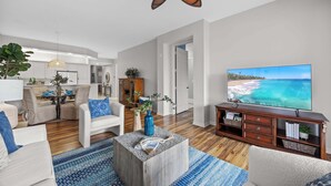 Ein geräumiges Wohnzimmer mit großem Fernseher, Holzboden und Deckenventilator.